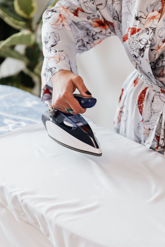 From above crop anonymous female wearing elegant silk robe ironing white fabric on blue ironing board by using steaming iron against white wall and green plants